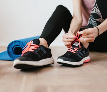 Crop sportswoman in sportswear tying sneakers while sitting on wooden floor with sport mat and dumbbells before exercising indoors