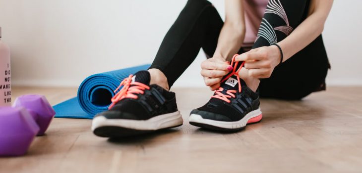 Crop sportswoman in sportswear tying sneakers while sitting on wooden floor with sport mat and dumbbells before exercising indoors