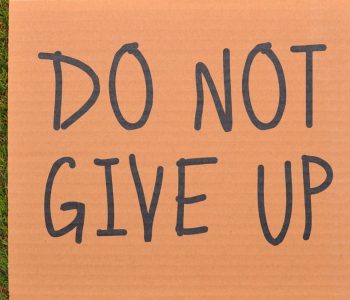 Cardboard sign reading 'Do Not Give Up' placed on green turf, symbolizing perseverance.