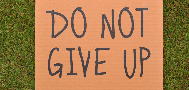 Cardboard sign reading 'Do Not Give Up' placed on green turf, symbolizing perseverance.