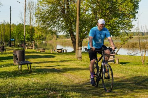 Adult man cycling along a lakeside path in a sunny park setting, surrounded by greenery and benches.