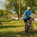 Adult man cycling along a lakeside path in a sunny park setting, surrounded by greenery and benches.